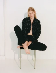 Model Bodine Van Galen seated barefoot on a metal chair wearing a black sequin jacket and black trousers against a white studio wall.