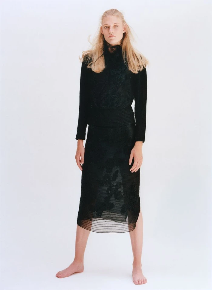 Model Bodine Van Galen wearing a black long-sleeved top and midi skirt against a seamless white background.