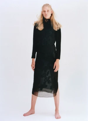 Model Bodine Van Galen wearing a black long-sleeved top and midi skirt against a seamless white background.