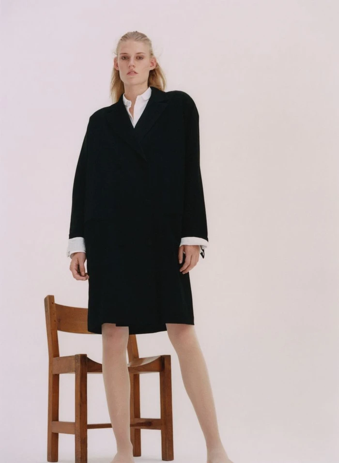 Model wearing a black coat and white shirt standing beside a wooden chair against a plain light background
