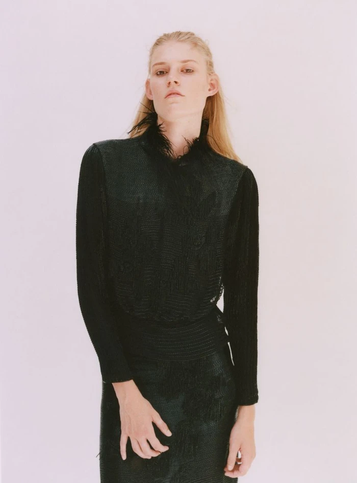 Model Bodine Van Galen in a dark textured garment with feather collar against a plain background