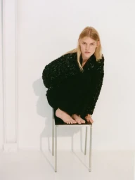 Model Bodine Van Galen crouching barefoot on a metal chair in a textured dark jacket against a plain white studio background.