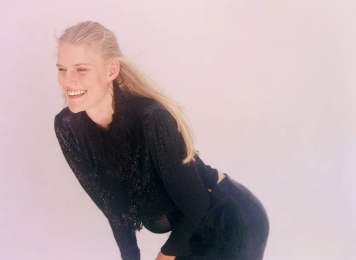 Model Bodine Van Galen smiling in a black textured top against a plain light background photographed by Alice Neale.