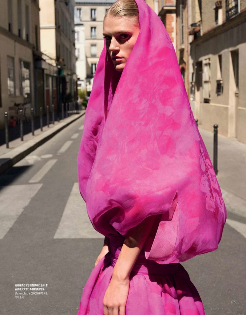 Model in a bright pink hooded garment standing on a sunlit city street for Vogue China