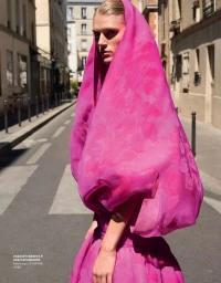 Model in a bright pink hooded garment standing on a sunlit city street for Vogue China