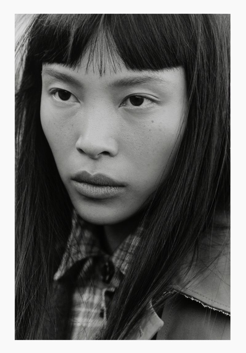 Black and white close-up portrait of an East Asian woman with blunt bangs wearing a plaid collared shirt and jacket.
