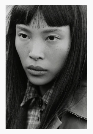 Black and white close-up portrait of an East Asian woman with blunt bangs wearing a plaid collared shirt and jacket.