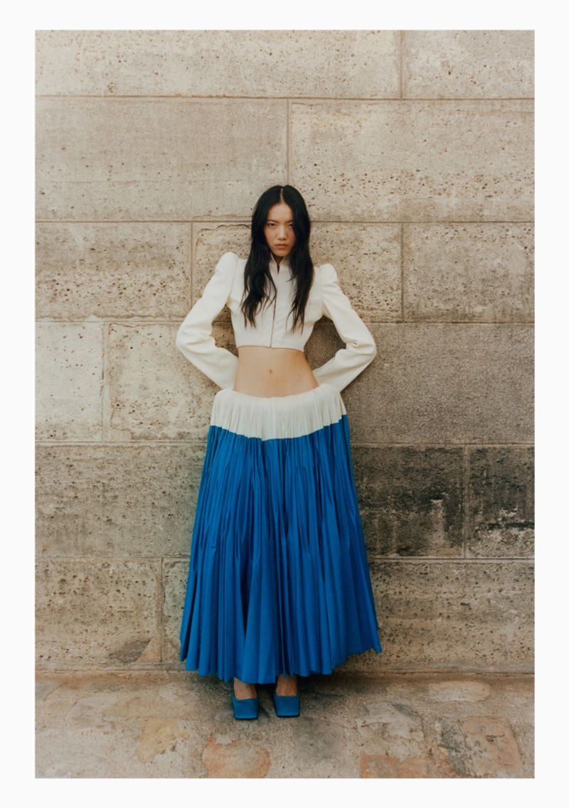 Fashion shot of an Asian woman posing against a stone wall, wearing a white jacket and a long blue and white skirt with blue shoes.