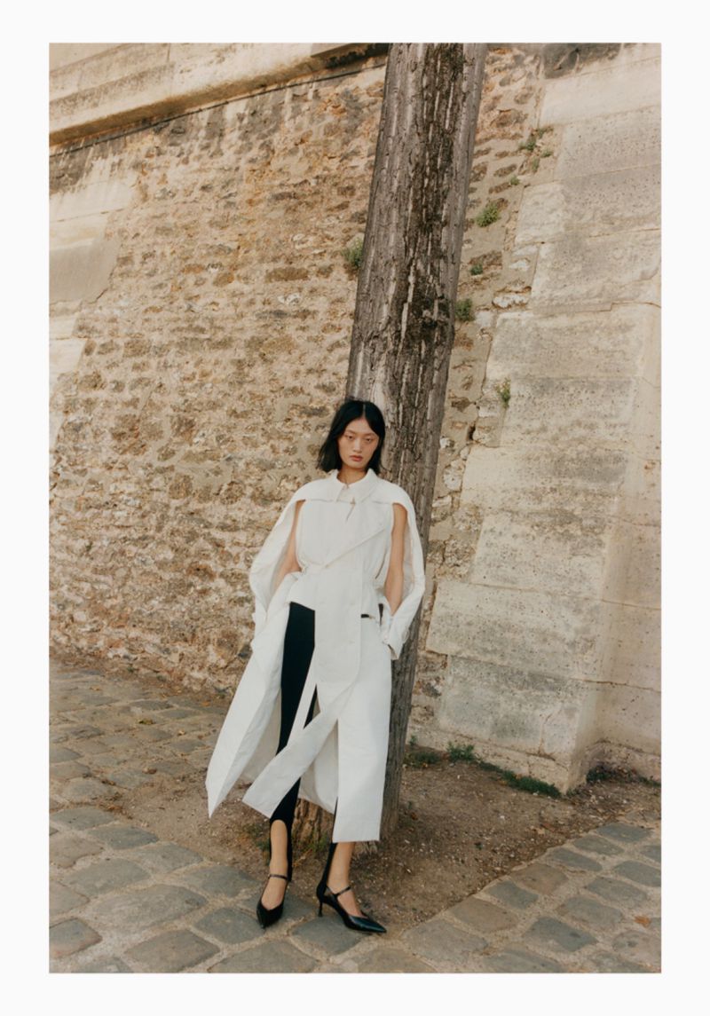 A fashionable woman in a long white coat poses outdoors near a stone wall and wooden post wearing black shoes, capturing a blend of style and setting.
