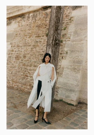 A fashionable woman in a long white coat poses outdoors near a stone wall and wooden post wearing black shoes, capturing a blend of style and setting.