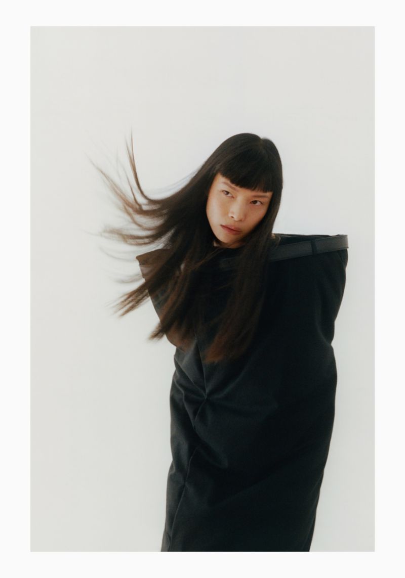 An Asian woman with long black hair poses in a black outfit against a white background, exuding a modern, high-fashion aesthetic in studio lighting.