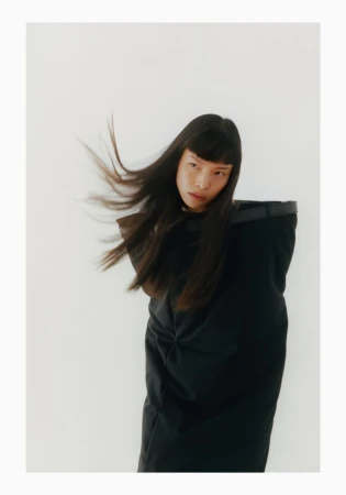 An Asian woman with long black hair poses in a black outfit against a white background, exuding a modern, high-fashion aesthetic in studio lighting.