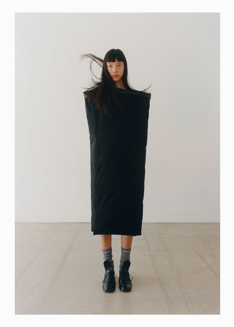 An Asian fashion model is posing in a black outfit in a studio with a white background, wearing black shoes and grey socks. 