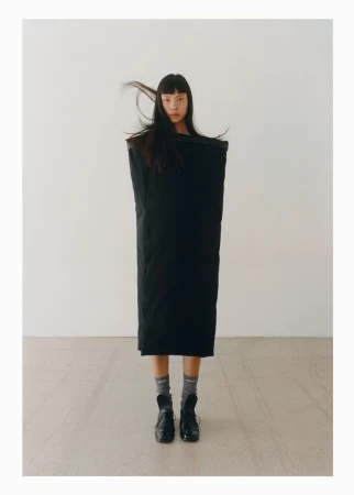 An Asian fashion model is posing in a black outfit in a studio with a white background, wearing black shoes and grey socks. 