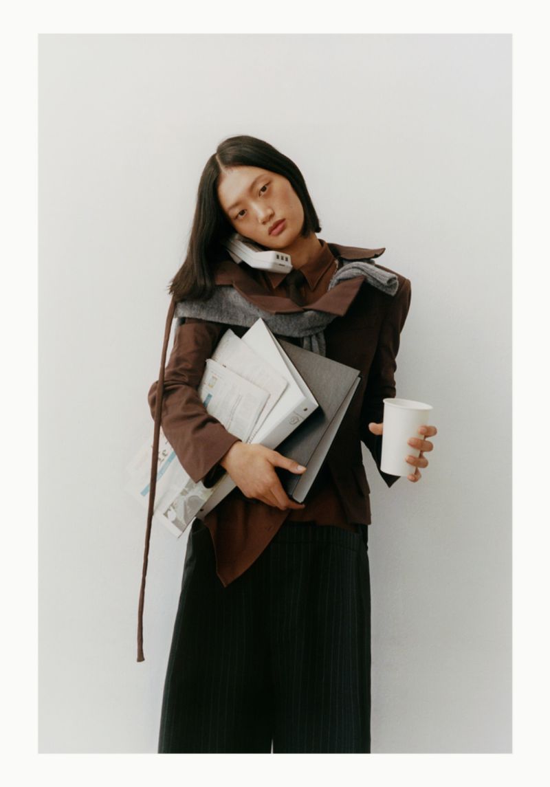 Adult woman in a brown jacket talking on a landline phone while holding a newspaper, laptop, and white cup against a plain white background.