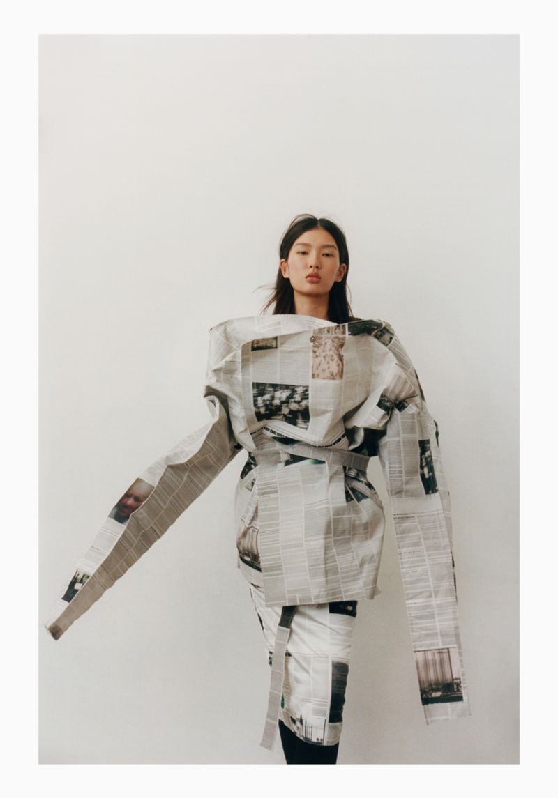 Fashionable adult woman wrapped in newspapers, striking a pose indoors. The image features a unique, editorial style with a white backdrop.