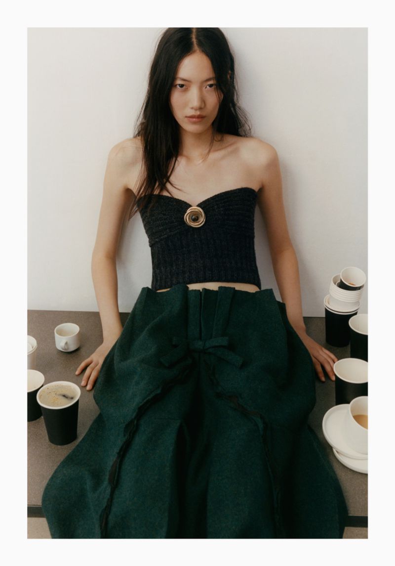 Young woman in a black tube top and green skirt posing indoors with coffee cups and plates scattered on a gray surface.
