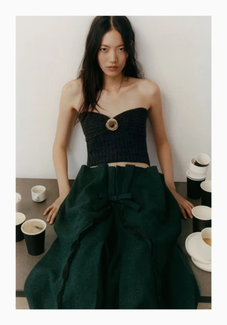 Young woman in a black tube top and green skirt posing indoors with coffee cups and plates scattered on a gray surface. 