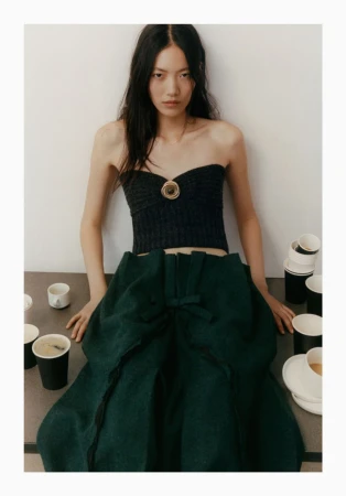 Young woman in a black tube top and green skirt posing indoors with coffee cups and plates scattered on a gray surface. 