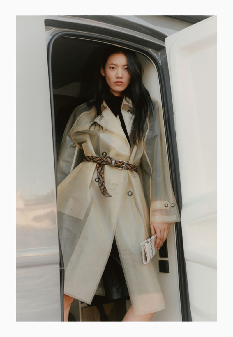 Asian woman in a clear trench coat and brown belt standing in the doorway of an RV with a neutral color palette and fashion styling.