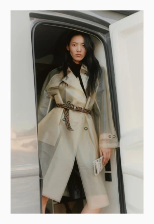 Asian woman in a clear trench coat and brown belt standing in the doorway of an RV with a neutral color palette and fashion styling.
