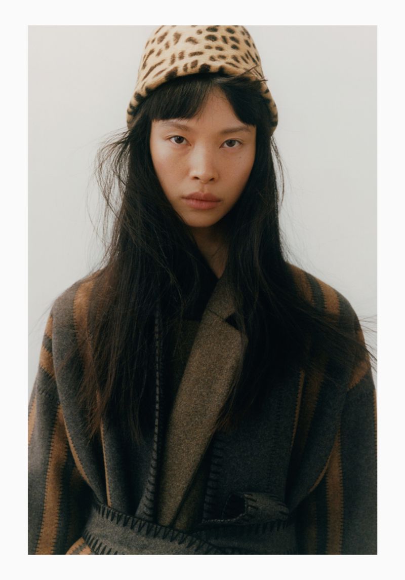 Headshot of an adult female model with long dark hair wearing a leopard print hat and striped wool coat in a studio setting.
