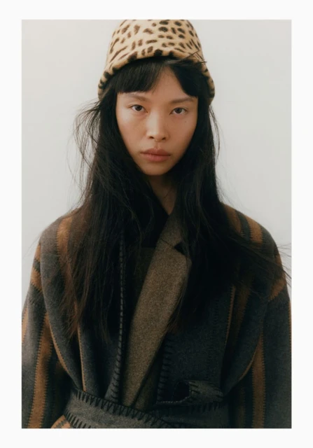 Headshot of an adult female model with long dark hair wearing a leopard print hat and striped wool coat in a studio setting.