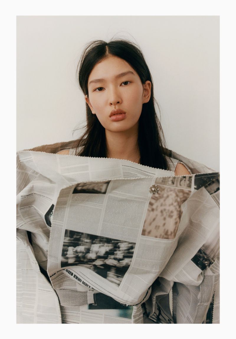 Asian woman gazes at the camera, draped in a newspaper-print garment against a plain backdrop, soft lighting, and medium shot framing.