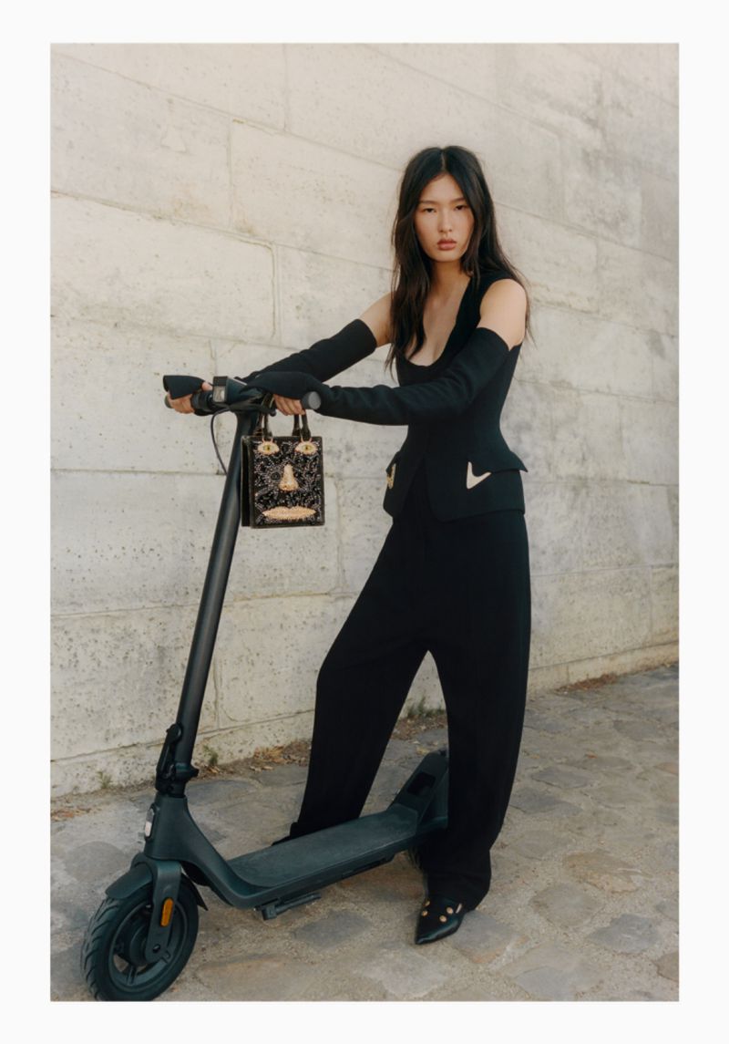 Adult Asian woman in sleeveless top and wide-leg pants posing on electric scooter with designer purse against a textured wall outdoors.