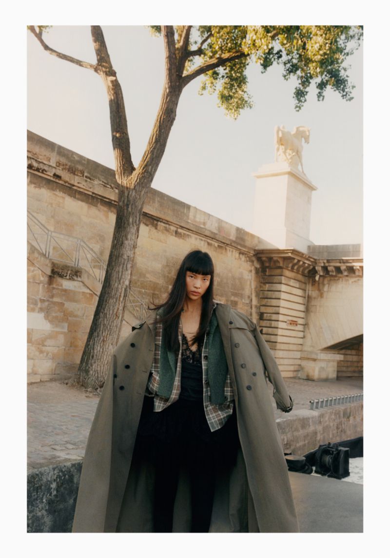 Asian woman in a trench coat standing by a stone bridge with a statue and tree; camera with a lens near the water's edge.