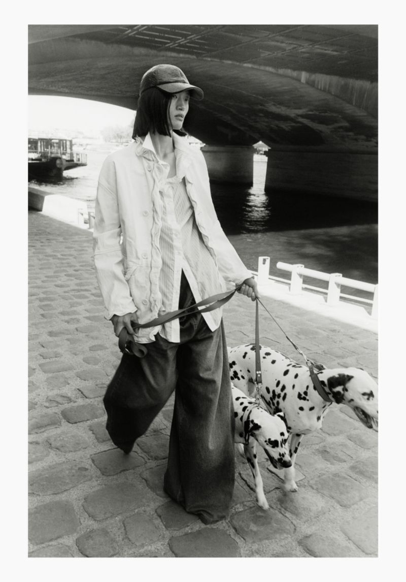 Black and white photograph of an East Asian model walking two Dalmatians on leashes along a cobblestone path under a large bridge.