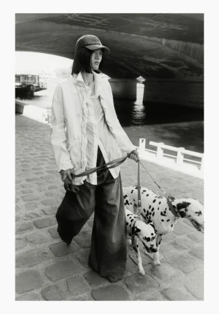 Black and white photograph of an East Asian model walking two Dalmatians on leashes along a cobblestone path under a large bridge.