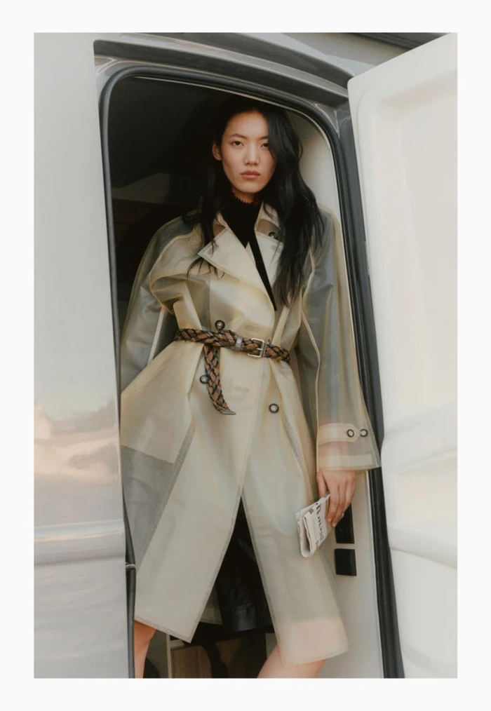 East Asian woman model wearing a translucent belted trench coat over black clothing, standing in the doorway of a white van.