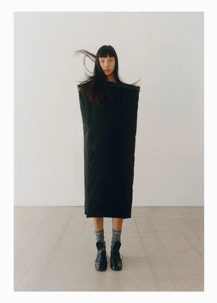 Model wearing a structured black dress and dark shoes with long hair blowing in front of a neutral background