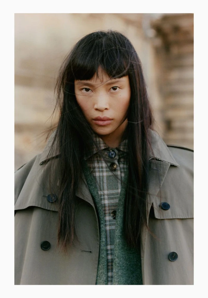 Portrait of a model with dark hair wearing an olive trench coat and plaid shirt outdoors.