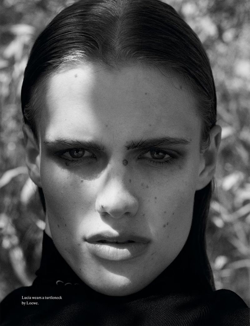 Close-up portrait of a model with dark slicked-back hair wearing a black turtleneck against an outdoor foliage background