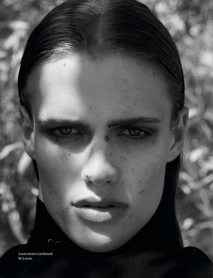 Close-up portrait of a model with dark slicked-back hair wearing a black turtleneck against an outdoor foliage background