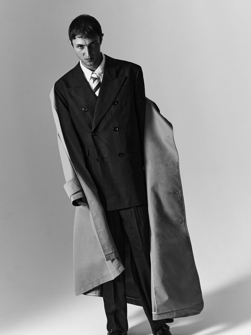 Anwar Hadid wearing a dark double-breasted suit and a long trench coat draped over his shoulders against a light studio backdrop.