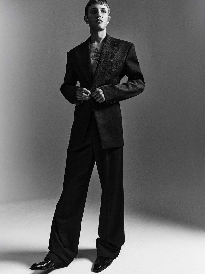Anwar Hadid wearing a black tailored suit and dark shoes standing against a grey seamless studio backdrop