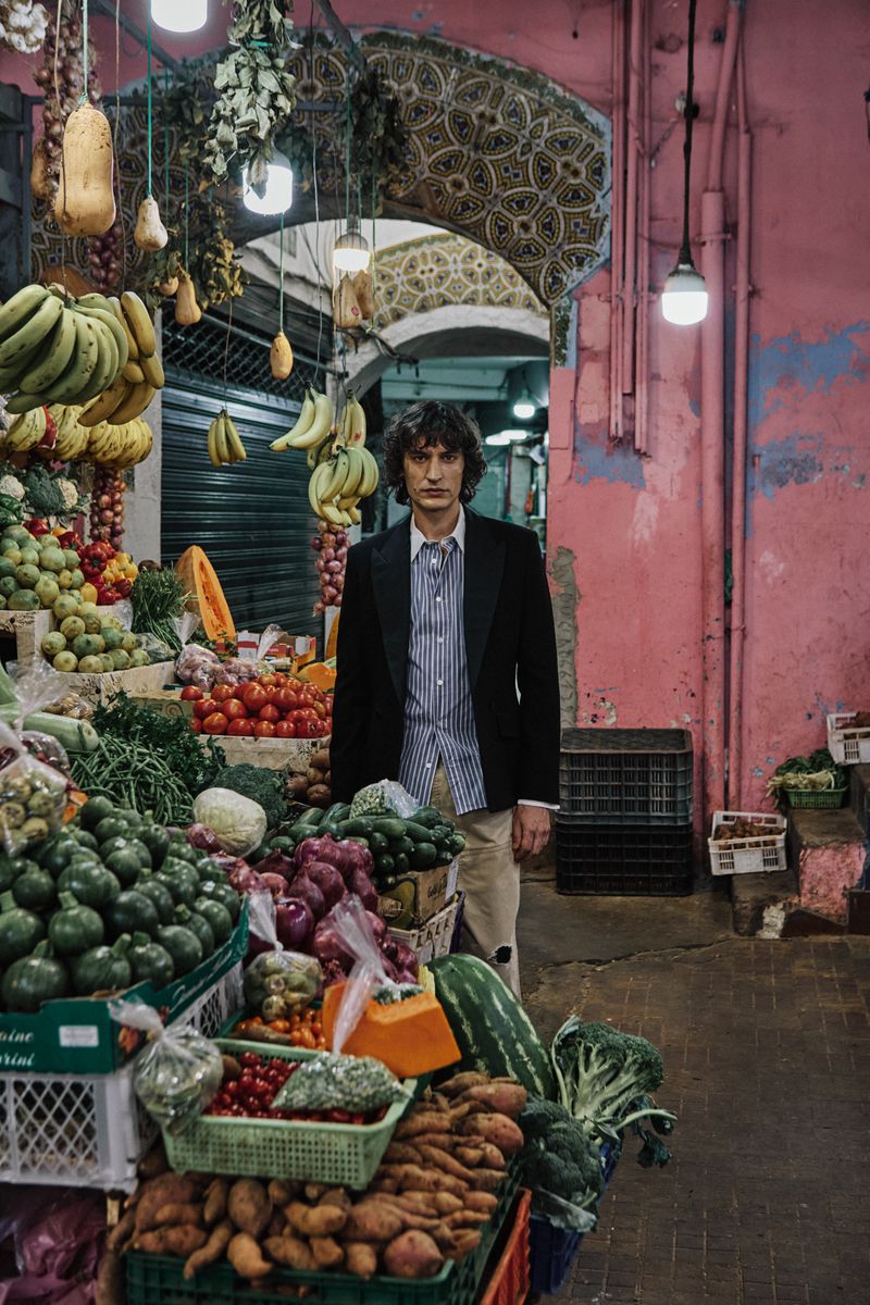 Man standing in a bustling market aisle filled with hanging produce and vibrant fruit and vegetable stalls.
