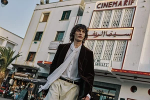 A man in a velvet blazer and white shirt stands outdoors in front of a white building with the sign CINEMA RIF.