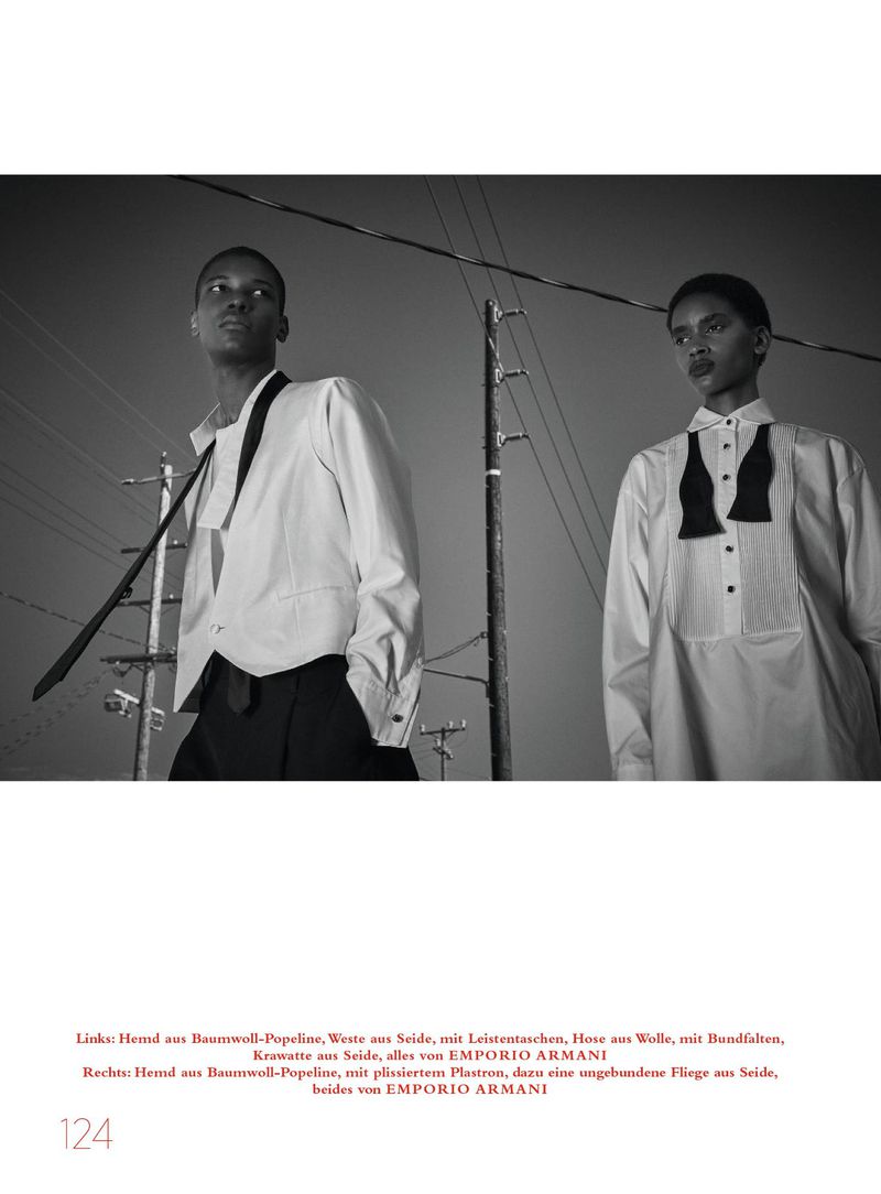 Two models wearing white shirts and dark trousers pose outdoors against utility poles and a pale sky in a black-and-white editorial image