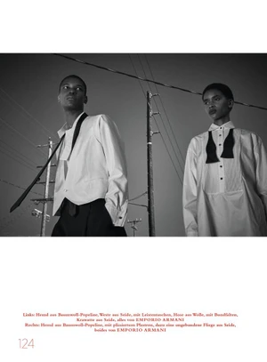 Two models wearing white shirts and dark trousers pose outdoors against utility poles and a pale sky in a black-and-white editorial image