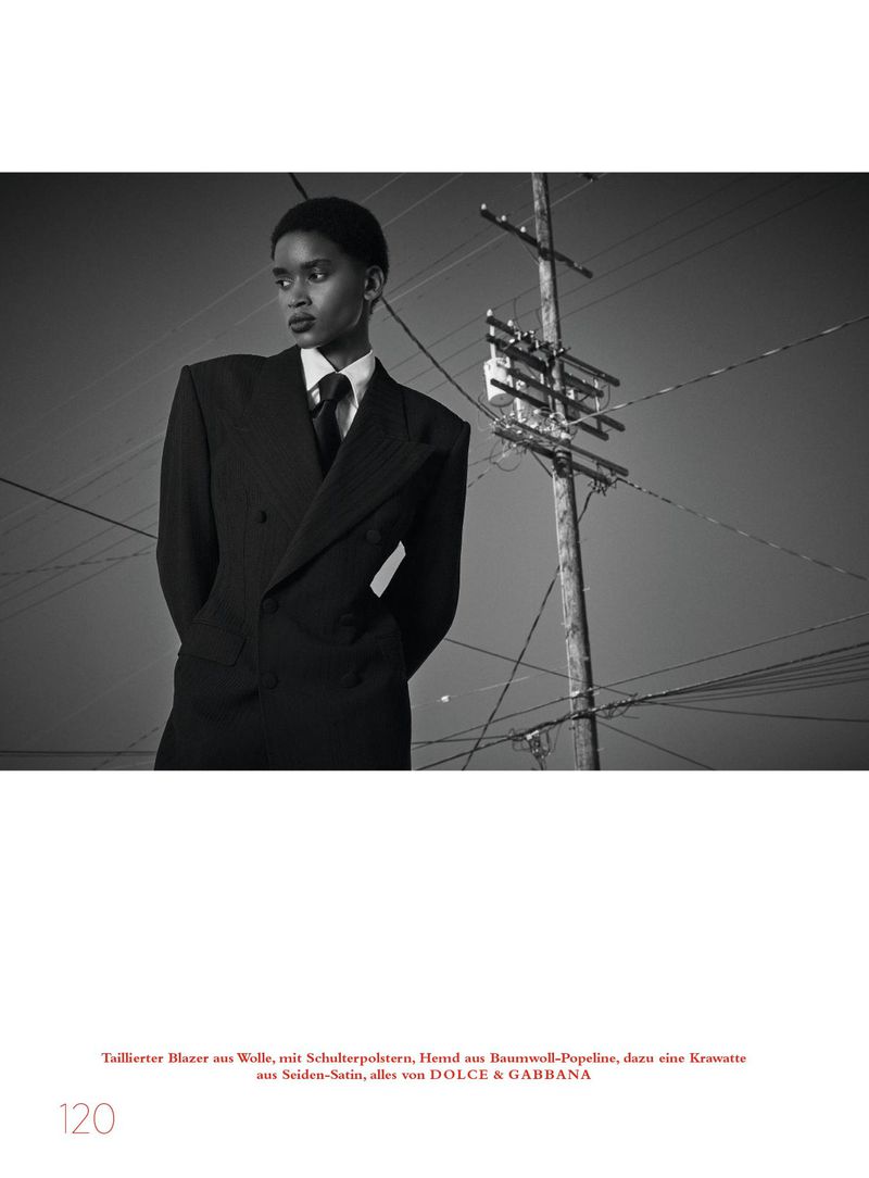 Model in oversized dark blazer and shirt with tie posing outdoors near a utility pole for Harper's Bazaar Germany.