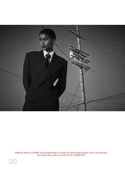 Model in oversized dark blazer and shirt with tie posing outdoors near a utility pole for Harper's Bazaar Germany.