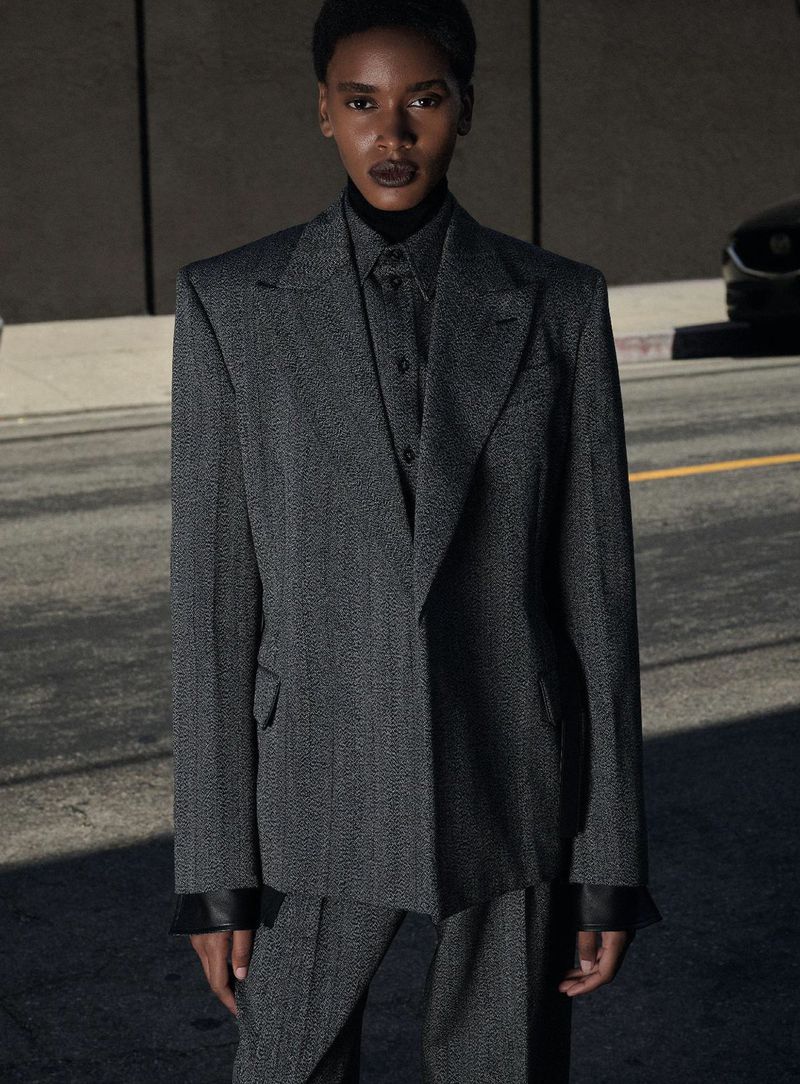 Model in a grey tailored suit jacket standing on a street with a car partially visible in the background.
