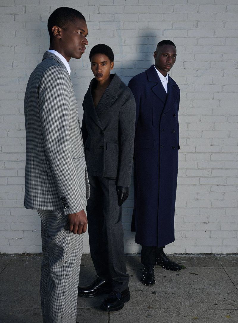 Three models wearing tailored suits and coats posing together outdoors against a white painted brick wall.