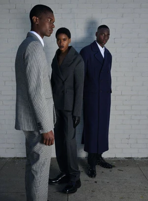 Three models wearing tailored suits and coats posing together outdoors against a white painted brick wall.