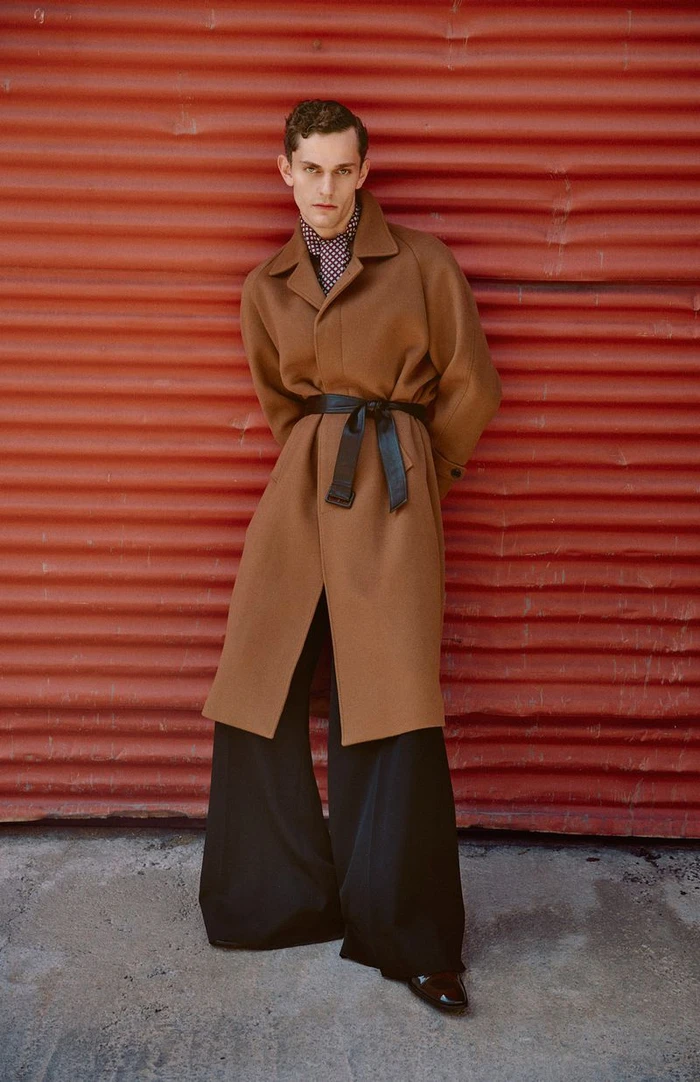 Male model wearing a long brown coat and black wide-leg trousers standing against a red corrugated wall