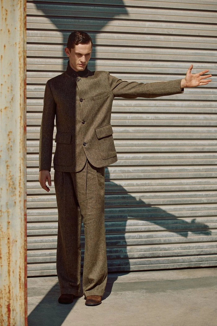 Model wearing a structured brown suit standing against a slatted metallic roller shutter door in an outdoor urban setting.
